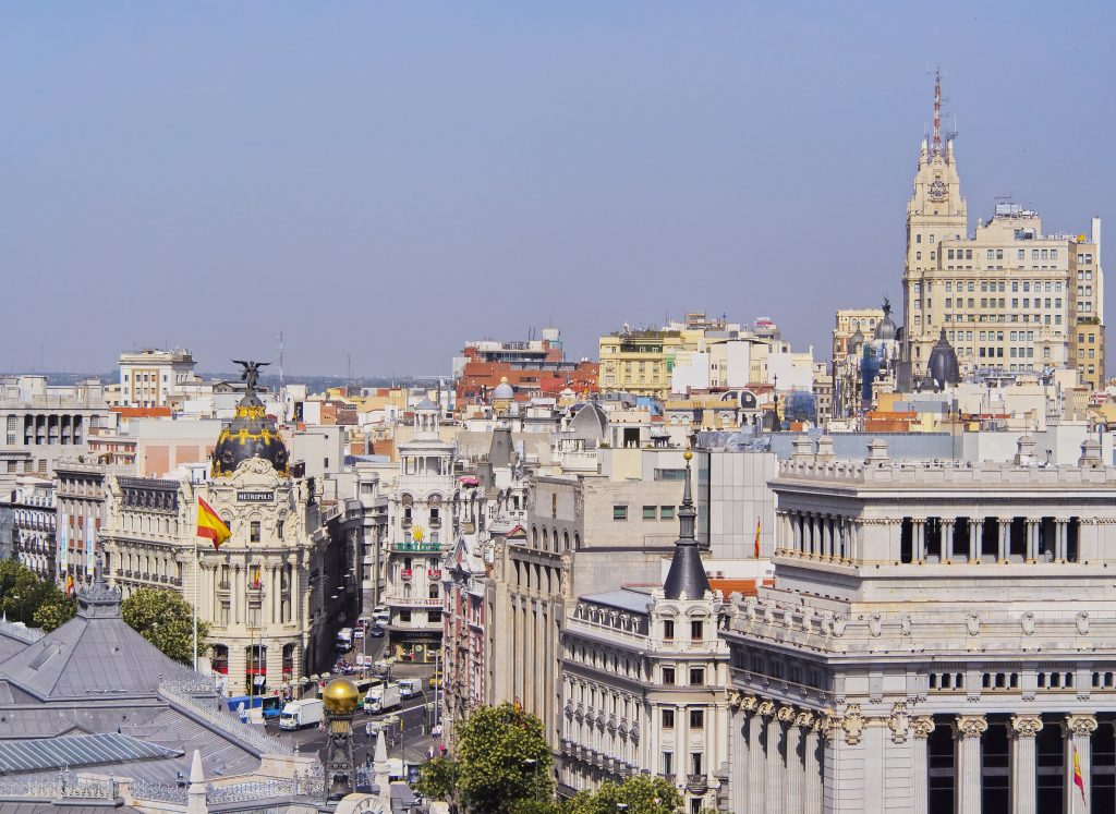 Classic Madrid cityscape showing ornate historic buildings, the Spanish flag, and bustling city streets under a clear blue sky.