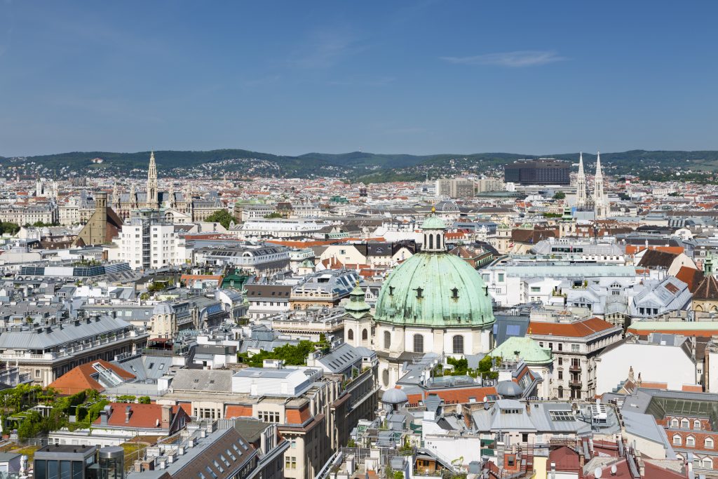 The cityscape of Vienna featuring historic architecture, the green dome of Peterskirche, and the distant hills of the Vienna Woods under a clear blue sky.