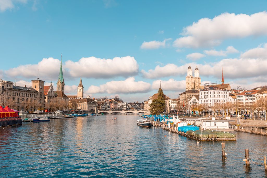 Scenic view of Zurich's old town along the riverfront under a blue sky with fluffy white clouds, with historic buildings and boats docked in the water.