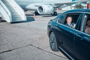 A high-end black luxury sedan provides VIP airside transfer service, with a male passenger in a business suit seen through the window as he gets into the car. A mobile airplane staircase and a large commercial jet with its engine visible are in the background on the airport tarmac.