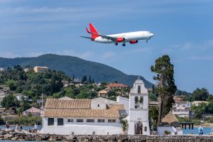 A commercial jetliner with distinct red engine and tail livery makes a low-altitude final approach, flying directly over the whitewashed Vlacherna Monastery in Corfu, Greece, with a hilly landscape and villas in the background.