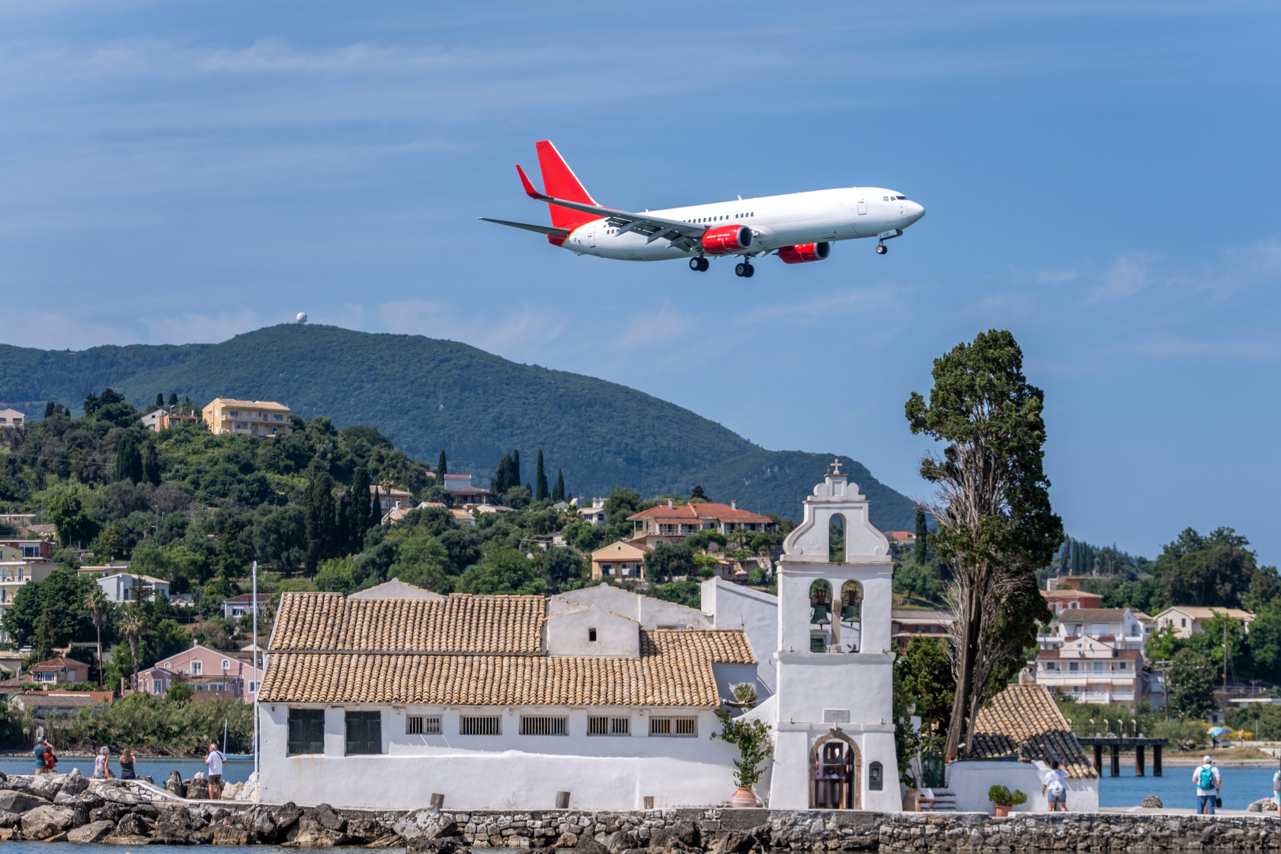 A commercial jetliner with distinct red engine and tail livery makes a low-altitude final approach, flying directly over the whitewashed Vlacherna Monastery in Corfu, Greece, with a hilly landscape and villas in the background.