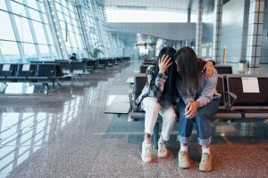 Two young women sitting in a bright airport waiting area. One is covering her face with her hand, appearing upset, while her friend sits beside her with a comforting arm around her shoulder.