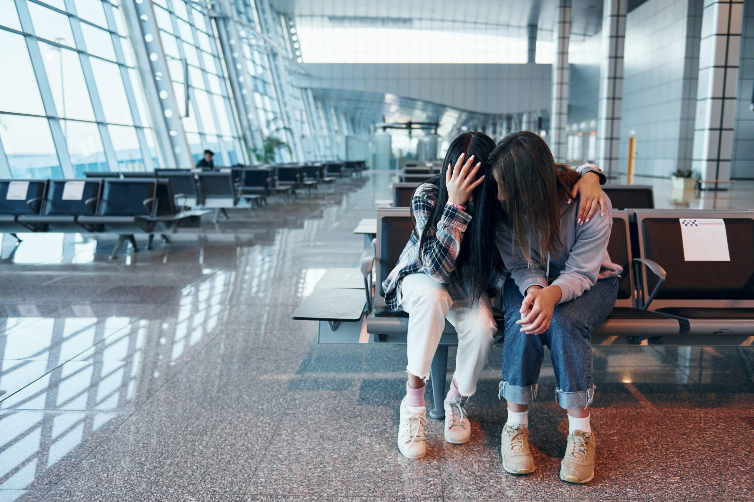 Two young women sitting in a bright airport waiting area. One is covering her face with her hand, appearing upset, while her friend sits beside her with a comforting arm around her shoulder.