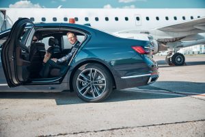 A well-dressed, cheerful man in a stylish suit steps out of a car during a transfer, heading toward a jet before takeoff.