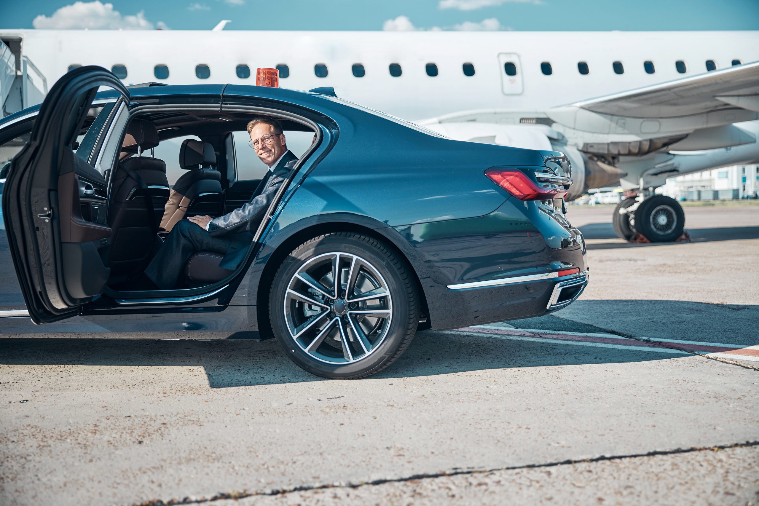 A well-dressed, cheerful man in a stylish suit steps out of a car during a transfer, heading toward a jet before takeoff.