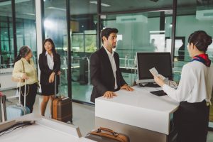 Alt text: A businessman presents his passport to an airline representative at the airport check-in counter while two women wait in line behind him, illustrating a real-world airport queue and customer service interaction.