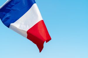 The national flag of France waving in the breeze against a bright blue sky, displaying the iconic blue, white, and red tricolor design.