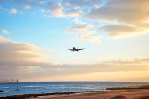 a commercial passenger jet is seen on final approach to ibiza airport, flying low over a coastal beach and the ocean at sunset, with an approach lighting tower visible in the water.