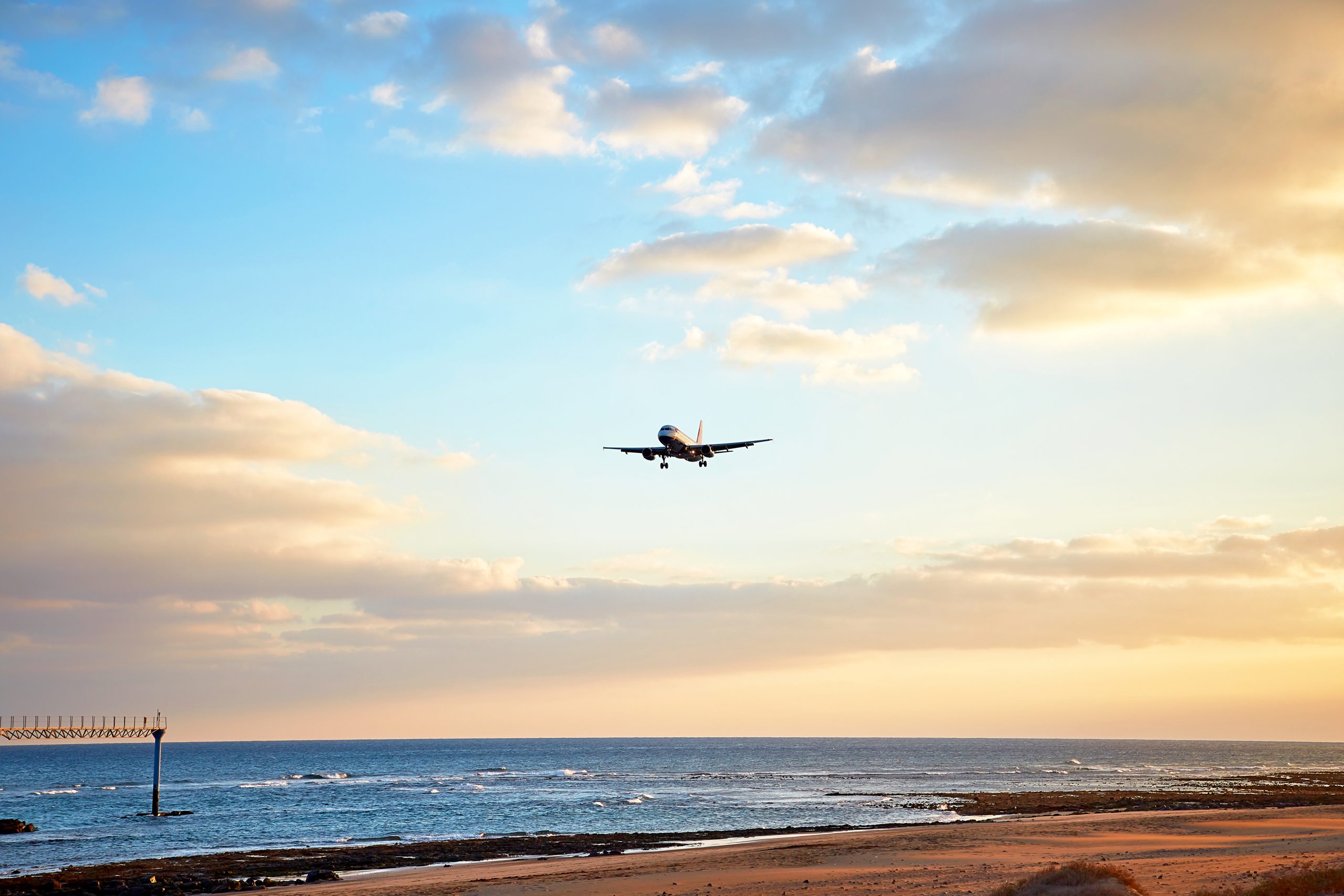 a commercial passenger jet is seen on final approach to ibiza airport, flying low over a coastal beach and the ocean at sunset, with an approach lighting tower visible in the water.