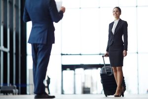 A cheerful professional woman wearing a dark skirt suit and white shirt stands in a brightly lit modern building lobby, holding the handle of a black rolling suitcase. In the out-of-focus foreground, a man in a blue suit stands with his back to the camera, appearing to greet her.