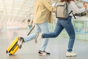 Two travelers walk through a bright airport concourse, pulling a yellow wheeled suitcase on a track.